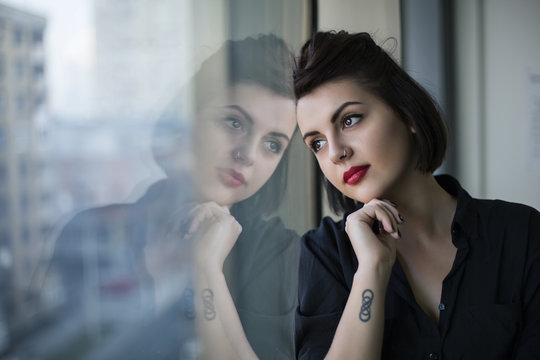 Young beautiful woman with black hair, piercing and a tattoo looking through a window