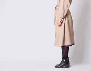 Woman's classic fall outfit with long beige trench coat, black tights and black ankle boots isolated on white background.