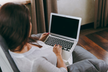 Fototapeta premium Beautiful young woman at home sitting on modern chair in front of window, relaxing in her living room and working with laptop. Close up. Back view. With place for text.