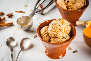 Ideas for autumn desserts, recipes from pumpkins. Pumpkin pie ice cream gelato in ceramic bowls, with maple syrup, pumpkin seeds, cinnamon and anise stars, on a white marble table. Copy space
