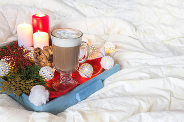 Breakfast with a cup of coffee, a biscuit on a tray on the bed on a white blanket in the winter morning decorated with Christmas decorations, spruce branches with luminous garlands, concept holiday