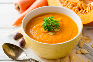 Pumpkin and carrot cream soup with pumpkin seeds and parsley in bowl on white wooden background.