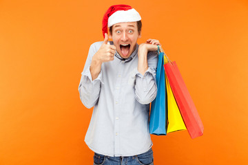 Young adult man  in christmas hat, holding  colorful bags after shopping. Thumbs up and tongue out.