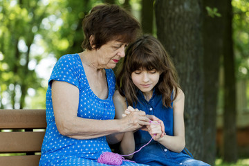 Grandmother teaches her granddaughter to crochet against green nature background.