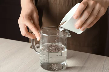 Fototapeten Woman adding chia seeds to water in measuring cup on table © Africa Studio