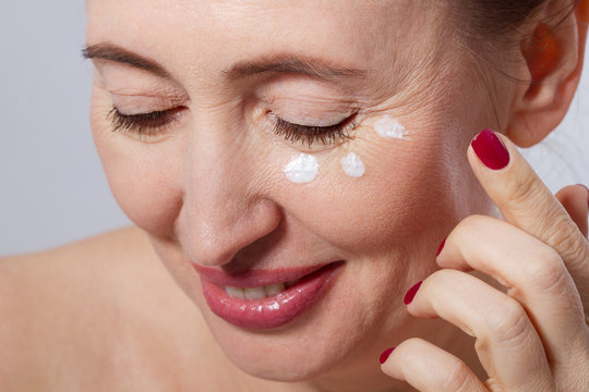 Beautiful middle aged woman applying cosmetic cream treatment on face isolated on grey background. Mock up and copy space. Cropped image