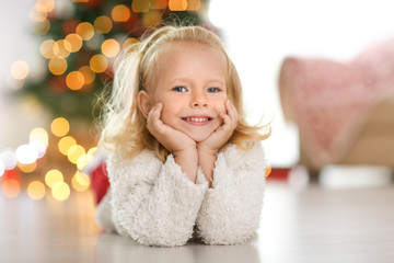 Fototapeta na wymiar Cute little girl lying on floor at home against defocused Christmas lights