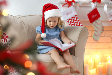 Obraz premium Cute little girl in Christmas hat reading book at home