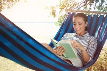 Young woman lying in hammock and reading book outdoors