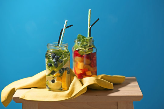 Mason jars of infused water with fruits and berries on wooden stool against color background
