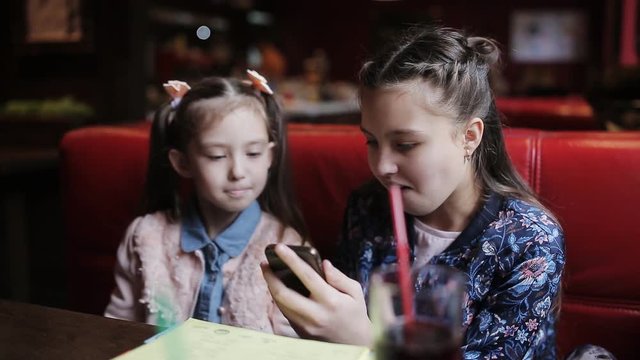 little girls sitting in a cafe, looking at the phone on the pictures and laughing.