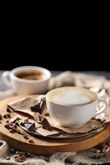 Composition with two glass of cappuccino and served with coffee beans and chocolate on wooden chopping board over black background