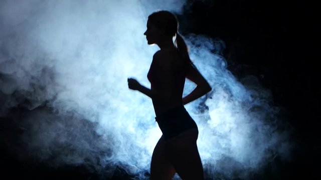 Athletic fitness girl running on a black background illuminated by the spotlight in the smoke. Silhouette