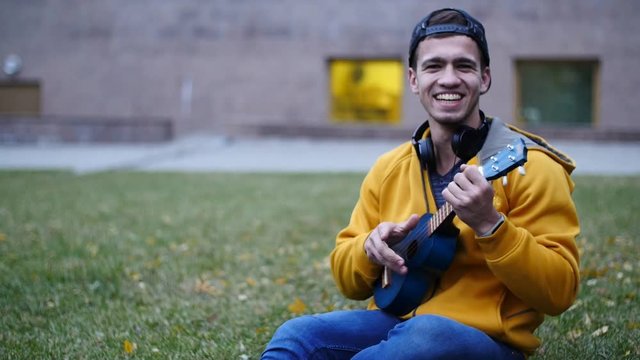 Attractive young man playing a ukulele sitting on grass