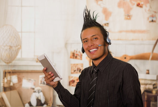 Close up of office punk man, wearing a suit with a crest, with headphones in his head and using his tablet in the office in a blurred background