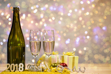 Bottle of champagne and glasses are placed together with a gift box and shiny balls. There is wooden texts "2018". Space on top right for your wording.