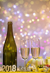Bottle of champagne and glasses are placed together with a gift box and shiny balls. There is wooden texts "2018". Space on top for your wording.