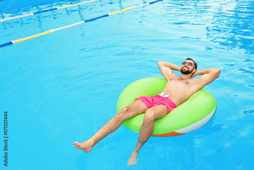 Wall mural Handsome young man with inflatable ring relaxing in blue swimming pool