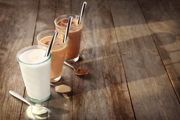 Glasses with different protein shakes and powders in spoons on wooden table
