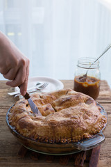 Traditional American apple pie in a glass pie dish. A female hand is cutting the pie with a knife. Caramel  sauce in the background. 