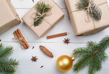 Fototapeta na wymiar Christmas background with gift boxes wrapped in kraft paper, fir tree branches, golden glass balls, pine cones, cinnamon sticks and stars anise on white wooden background. Flat lay, top view