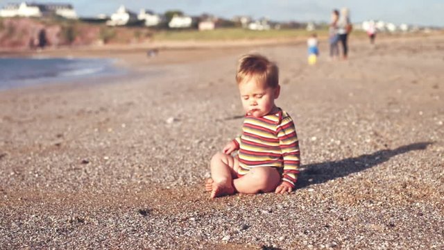 A cute little baby boy is sitting on the beach at sunset