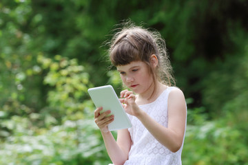 Little girl in white dress plays with tablet pc in green park at summer day