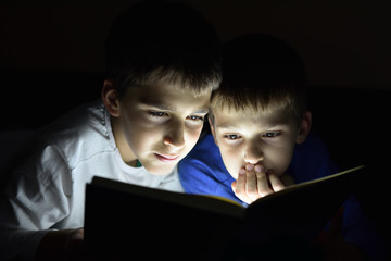 Two brothers reading a book hiding in the dark and using a torch light. Selective focus.