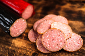 Pile of delicious smoked sausage sliced on a burnt wooden cutting board. Focus on front slice. Red and black sausages blurred in background.