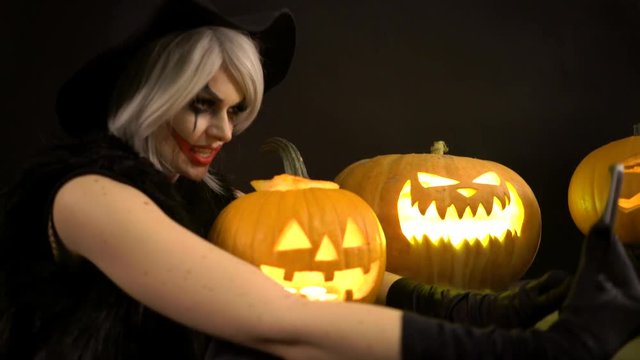 Scary beautiful girl witch celebrates halloween, makes selfie with funny glowing burning pumpkins in smoke. Woman with gray hair conjures, casts a spell. Gates of hell in fire