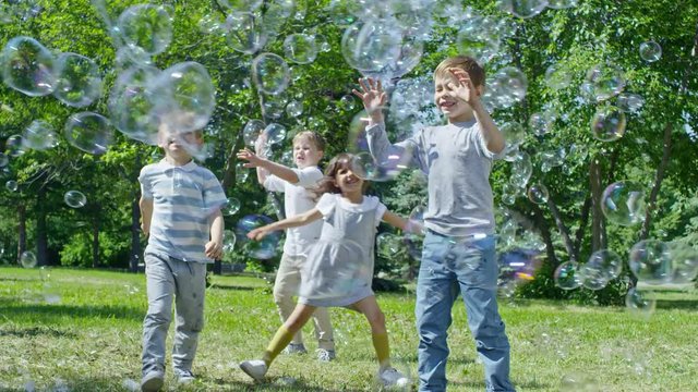 SLOWMO of little girl and three boys chasing and bursting large soap bubbles at sunny day in the park