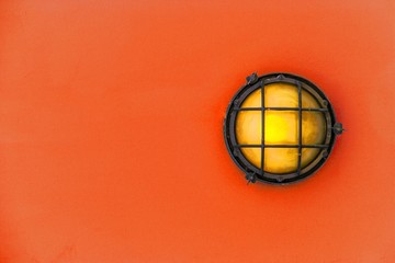 Yellow bulkhead light (ship deck lamp) surrounded by a metal rusted frame fixed to a painted red orange color wooden wall background. - 177251816