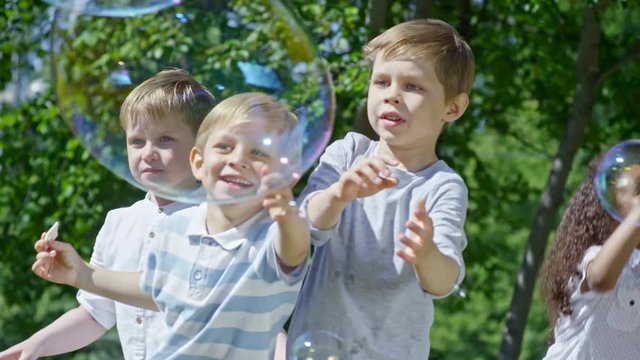 SLOWMO of three little boys trying to burst large soap bubble floating in the air while funny African girl jumping and playing behind them at warm summer day in park