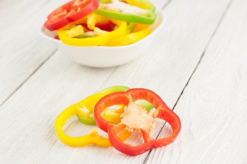 Two slices of colored red and yellow fresh ripe raw pepper beside heap of slices of pepper in ceramic bowl on old rural white wooden planks