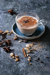 Glass cup of traditional indian masala chai tea on blue saucer with ingredients above. Cinnamon, cardamom, anise, sugar, black tea over dark texture background with space