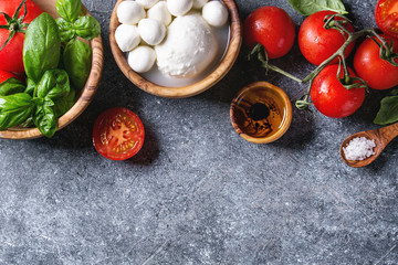 Ingredients for italian caprese salad. Mozzarella balls, buffalo, tomatoes, basil leaves, olive oil with vinegar, salt in olive wood bowls over gray texture background. Top view with copy space
