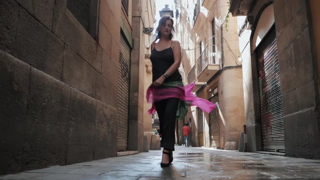 Front view of sexy elegant woman walking in the old european city. Attractive lady in long black dress, colorful scarf and heels in gothic quarter of Barcelona. Slow motion.