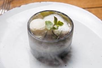 ice cream sweet dessert on a white plate on a wooden table in an Italian retor. Ingredients white ice cream with mint and cocoa in a transparent tequal glass with smoke.