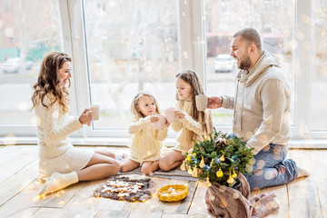 Happy family in white sweaters drinks cocoa by the panoramic window. Parents and daughters are...