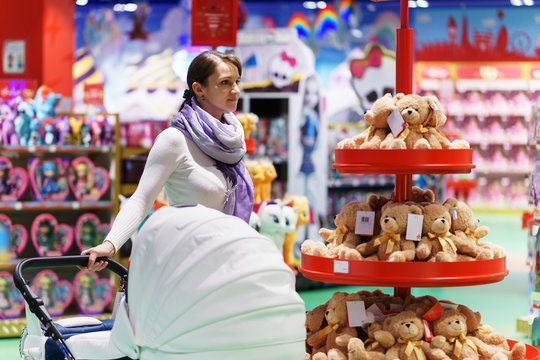 young caucasian moter is loking some toy at big supermarket for for her infant kid in white baby carriage