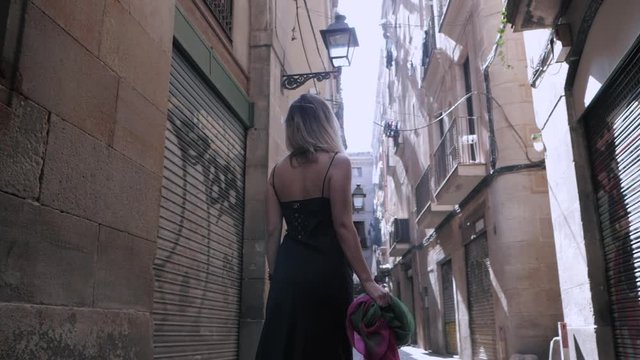 Rear view of sexy elegant woman walking in the old european city. Attractive lady in long black dress, colorful scarf and heels in gothic quarter of Barcelona. Slow motion.