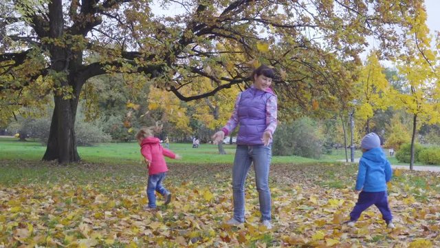 Happy family throws yellow leaves falling up. Mother with little son and daughter throwing leaves in Autumn in slow motion, smiling. Children playing in the autumn park foliage.