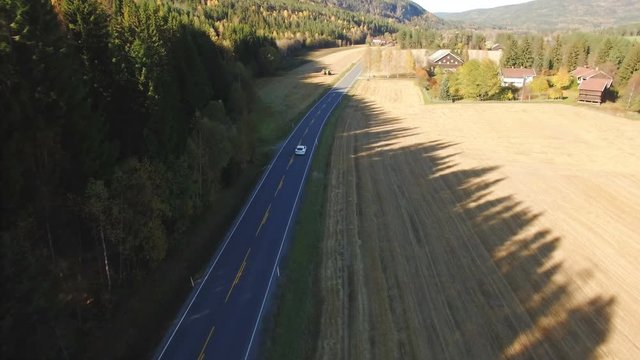 Modern luxury white electro car driving on empty country road among the woods and fields of autumn Norway. Top view aerial footage.