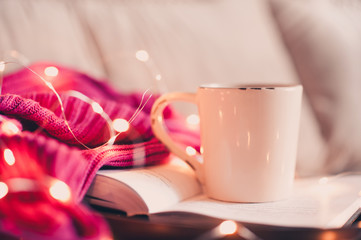 Cup of coffee staying on open book with pink knitted sweater and Christmas lights at background....