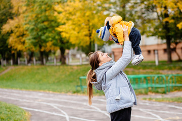 Happy sporty mother with infant son in warm clothers outdoors in autumn day. Young brunette mother lifting her son on stadium track - having fun enjoying motherhood. concept.