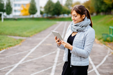 Young brunette sporty woman wiith tablet in hands dressed warm clothes and standing over stadium track background. Sport, lifestyle and multitacking concept.