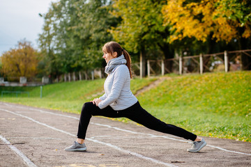 Young brunette sporty woman warming up - doing stertching exercises in track of stadium. Autumn,...