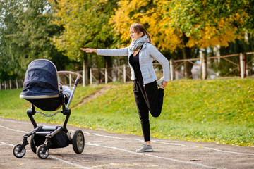 Young smiling sporty woman with baby stroller in warm clothes warming up - doing stertching...