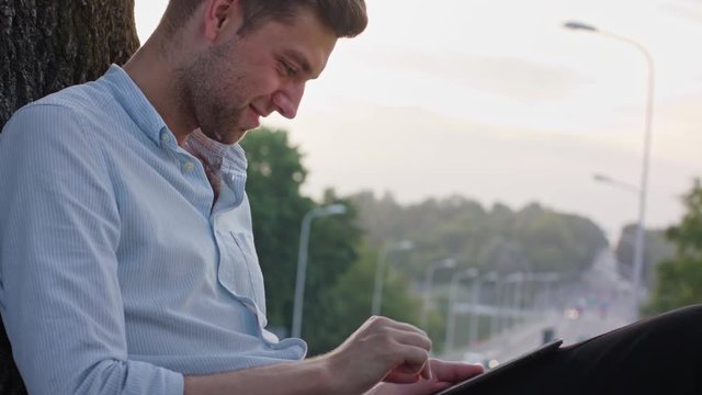 A young man using a tablet and sitting under the tree. Medium shot. Soft focus.