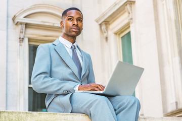 Way to success. Young African American College Student studying in New York, dressing formally in suit, tie, siting outside office building on campus, working on laptop computer, looking up, thinking